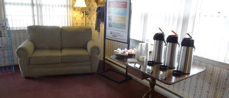 A living room with a sofa, a table with thermos flasks, a framed menu, and a trash bin.