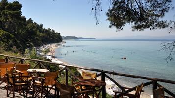 A seaside terrace with wooden chairs and tables, overlooking a beach and calm sea.