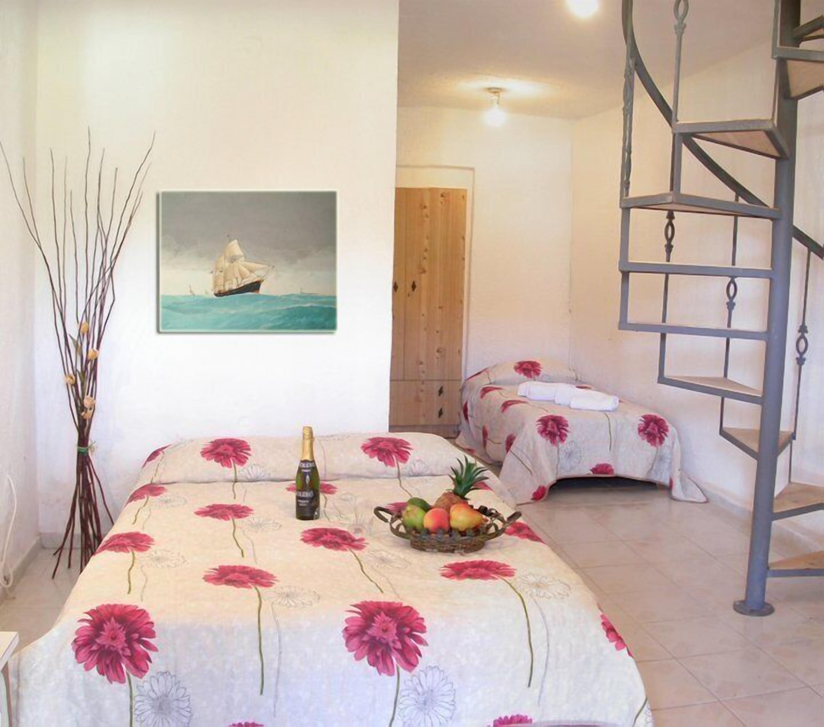 A room with two beds, a painting of a sailboat, a bottle of champagne, and a fruit basket.