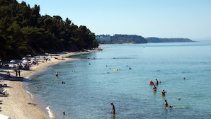 A beach with people swimming and sunbathing, a sandy shore, and a forested hillside. - Hotel Pighi (Kassandra)
