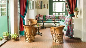 A room with a wooden ceiling, green-framed windows, a sofa with colorful cushions, wicker chairs, and a hanging lamp.