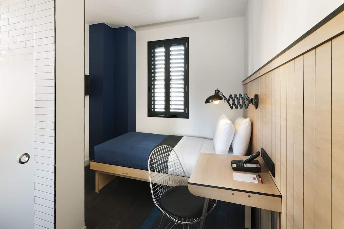 Une petite pièce comprenant un lit, un bureau, une chaise, une fenêtre avec des stores et une lampe fixée au mur.