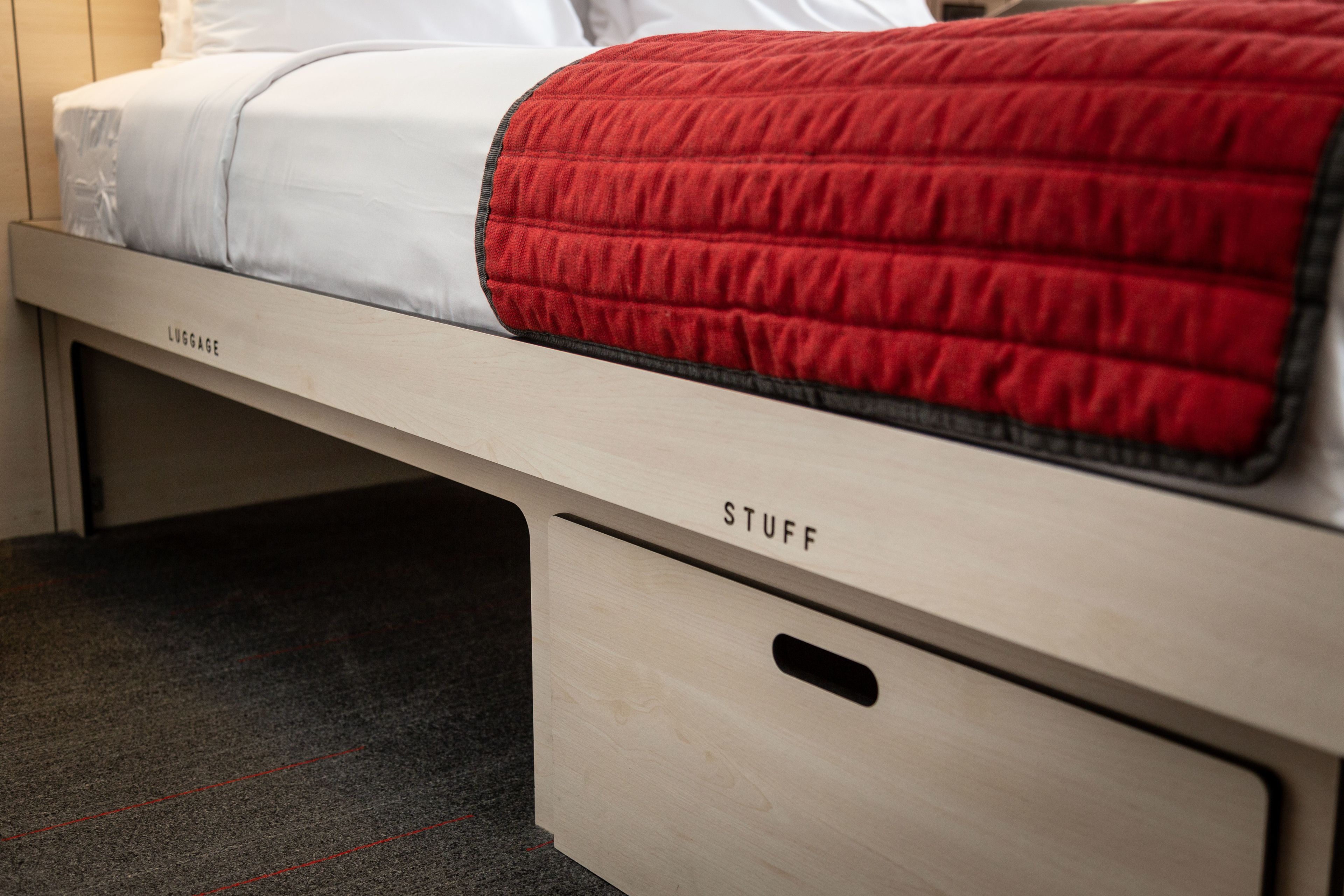 Un letto con materasso bianco e coperta rossa, un poggiatesta in legno con la scritta "LUGGAGE" e un cassetto con l'etichetta "STUFF".