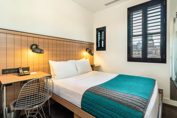 A hotel room with a bed, a desk with a chair, a telephone, and a window with blinds.