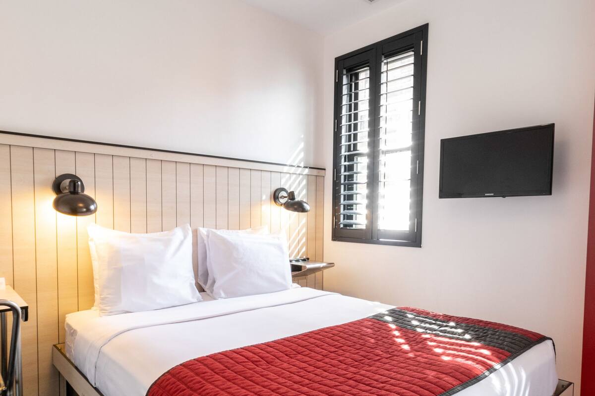 Un lit bien fait, avec une literie blanche et rouge, une lampe de chevet et un téléviseur fixé au mur.