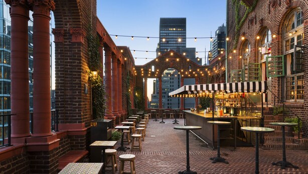 An outdoor patio with brick flooring, wooden tables and stools, and string lights.