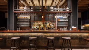 A bar area with a wooden counter, multiple stools, shelves stocked with bottles, and hanging glasses.
