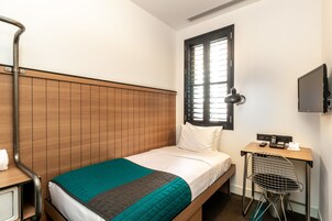 Een enkel bed met een houten hoofdbord, een bureau met stoel, een televisie en een raam met jaloezieën.