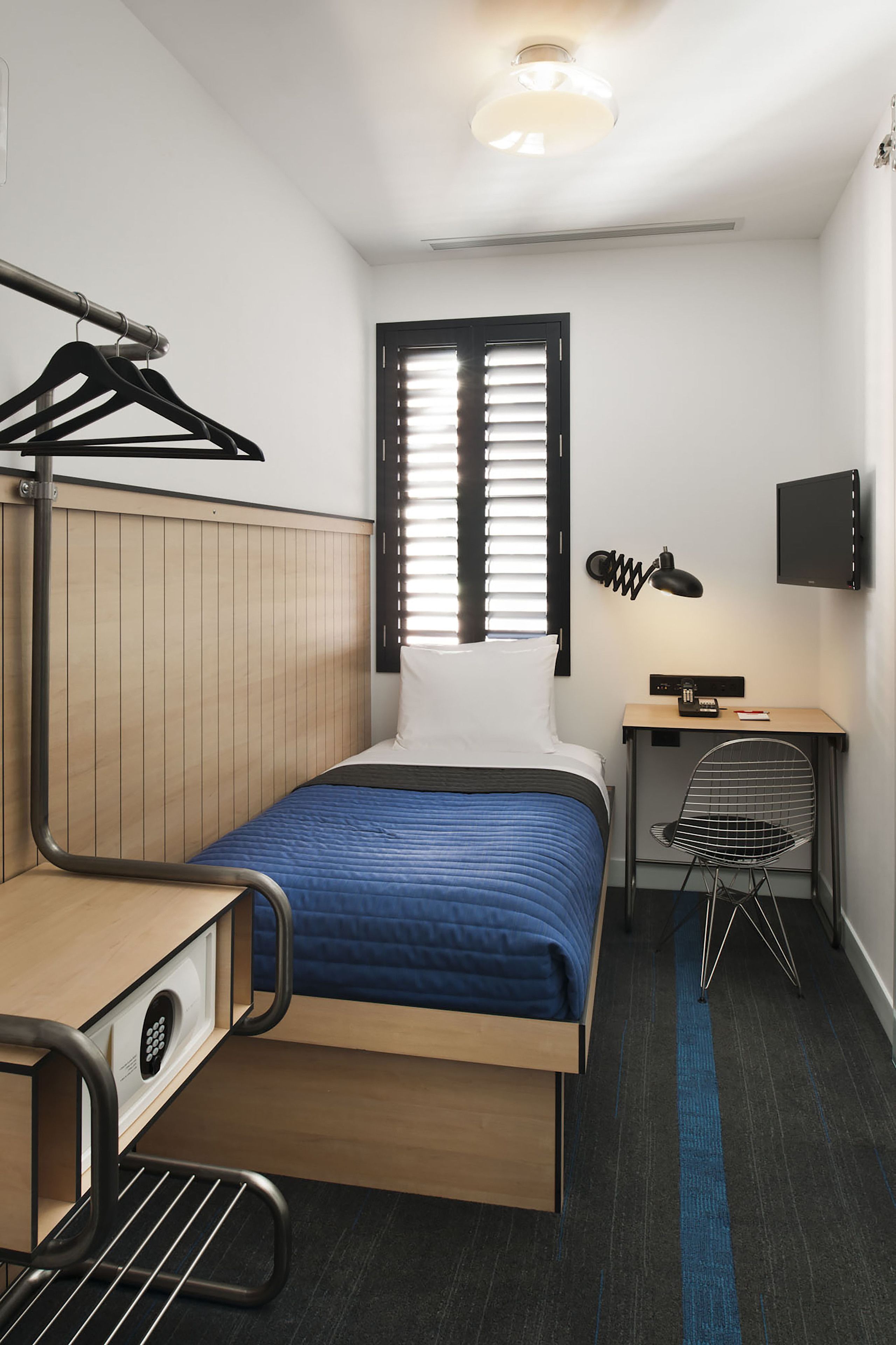 Quarto de hotel pequeno com cama, escrivaninha, cadeira e televisão fixada na parede.
