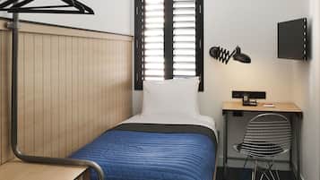 Quarto de hotel pequeno com cama, escrivaninha, cadeira e televisão fixada na parede.