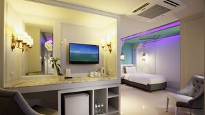 A modern hotel room with a large mirror, a flat-screen TV, a bed, a seating area, and a minibar. - The Verandah (Krabi)