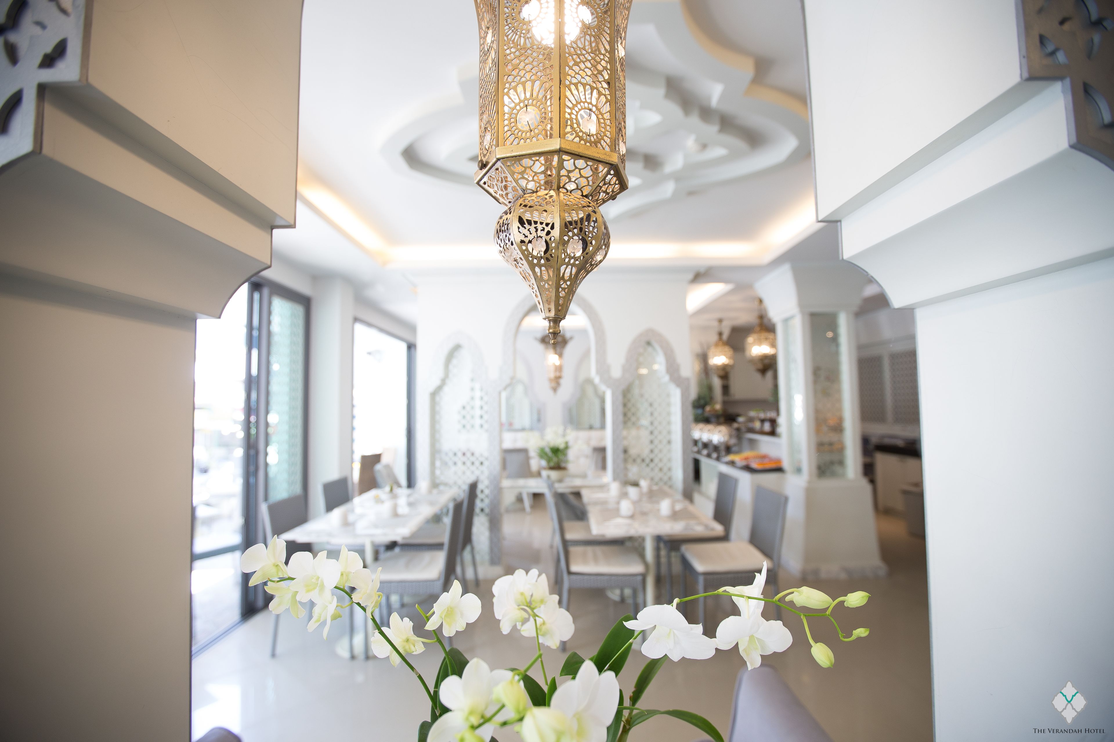 Un comedor con mesas y sillas blancas, una lámpara decorativa colgante y vista de la cocina al fondo.