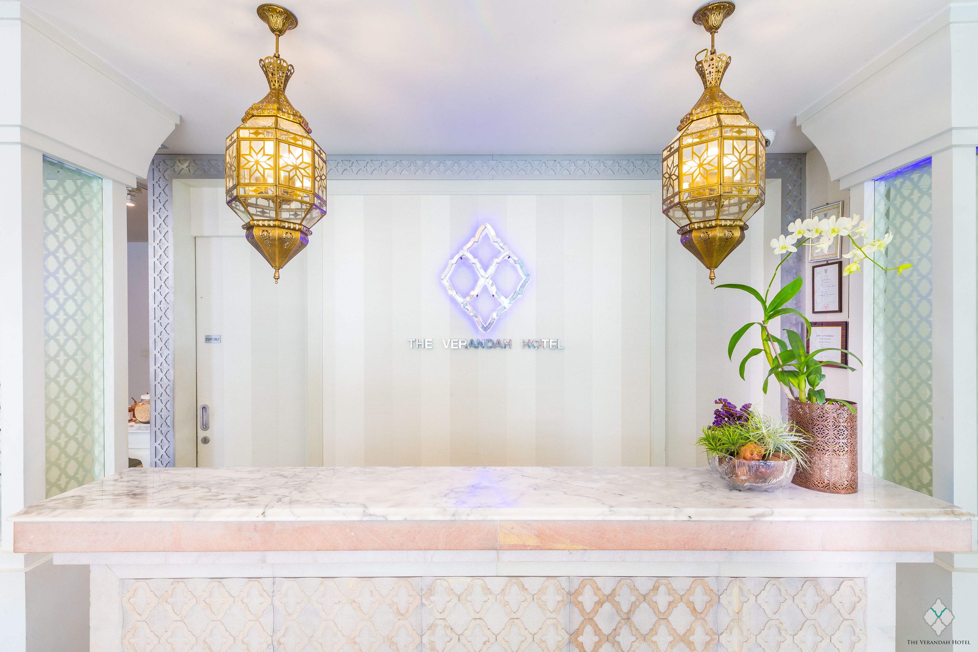 Uma área de recepção de hotel com balcão de mármore, duas luminárias pendentes ornamentadas, uma parede decorativa com padrão geométrico e uma placa com a inscrição 
