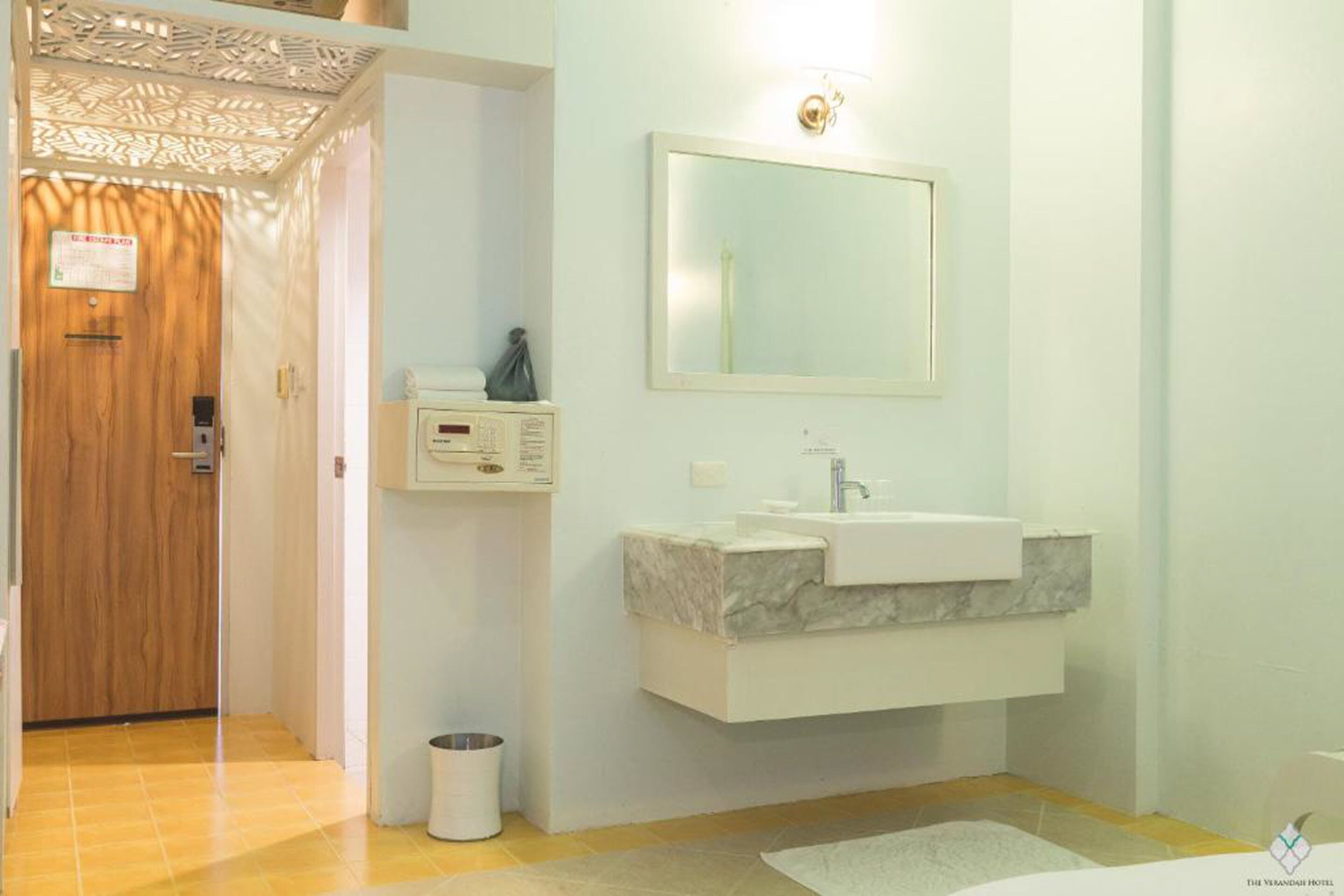 A modern bathroom with a wooden door, a white sink with a marble countertop, a mirror, and a small trash bin.
