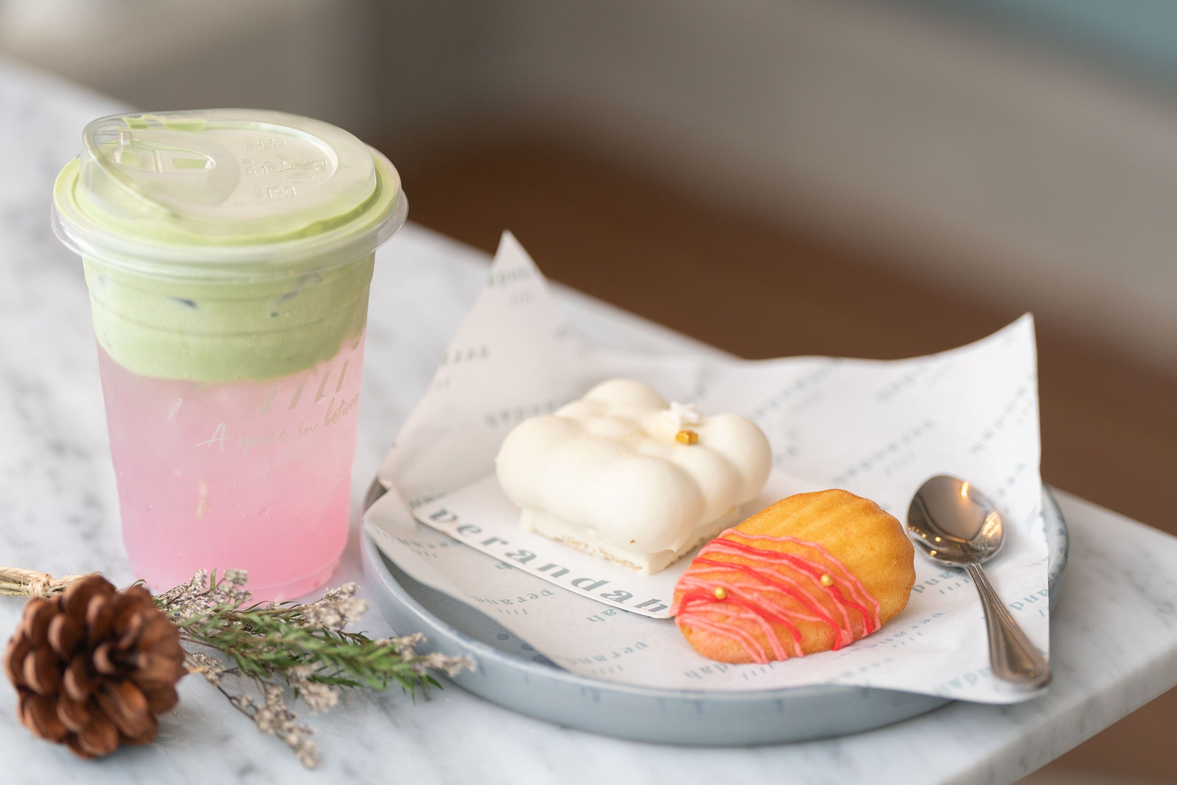 A dessert plate with a pastry and a spoon, a drink with a lid, and a pine cone on a marble surface.