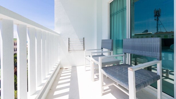 A balcony with white railings, two wicker chairs, and a large window with a view of a building and a communication tower. - The Verandah (Krabi)