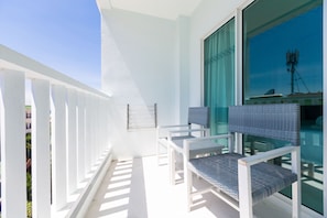 Un balcon avec des balustrades blanches, deux fauteuils en osier et une grande fenêtre donnant sur un immeuble et une tour de communication.