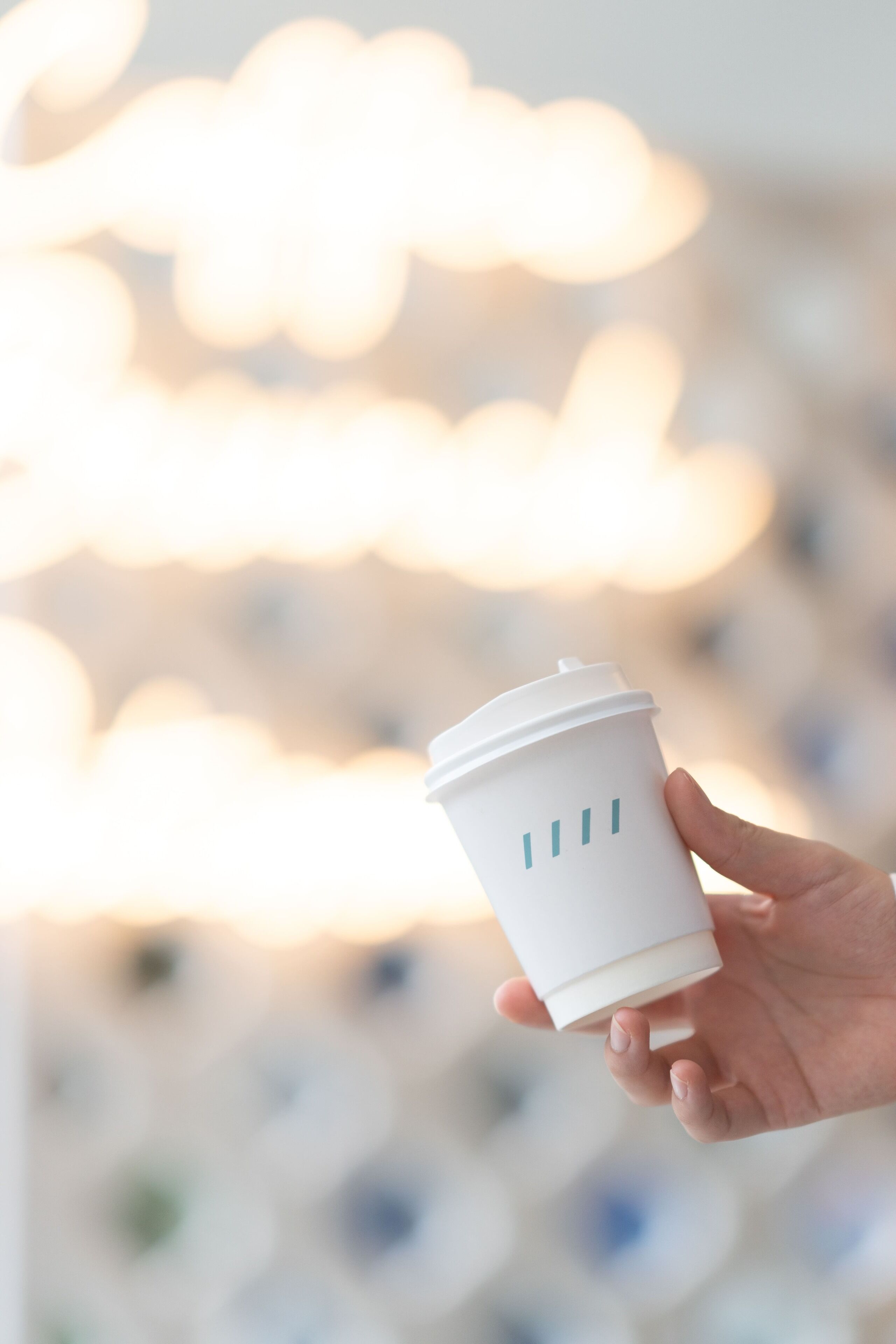 A hand holding a white disposable cup with a lid, set against a blurred background with bokeh light effects.