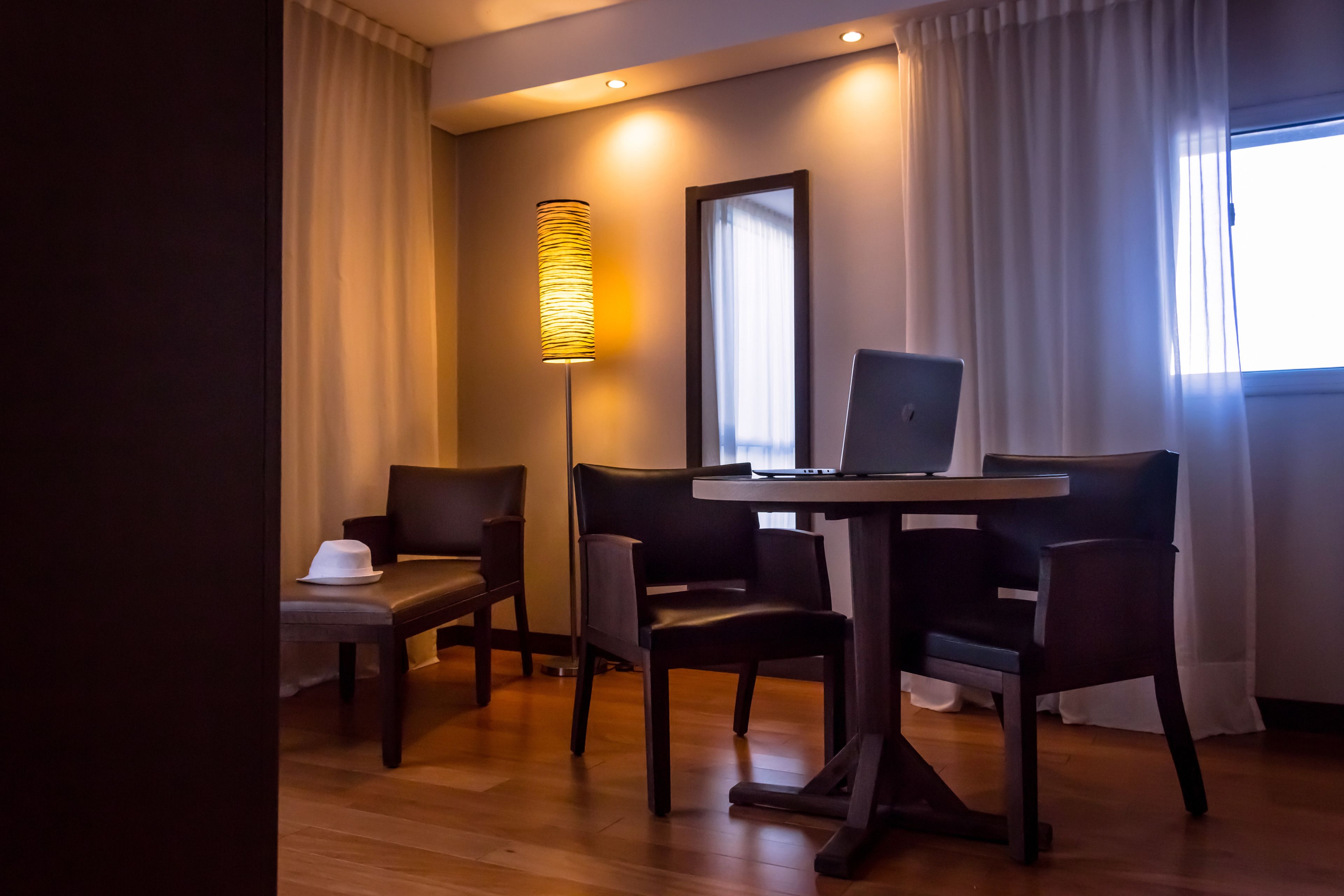 Habitación de hotel con escritorio, laptop, dos sillas, lámpara de pie y ventana con cortinas.