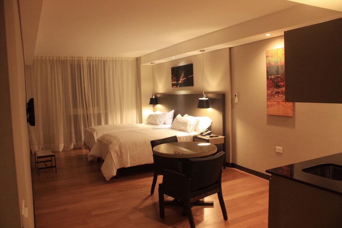 Una habitación de hotel con cama, mesita de noche, lámpara, silla y una pequeña mesa.