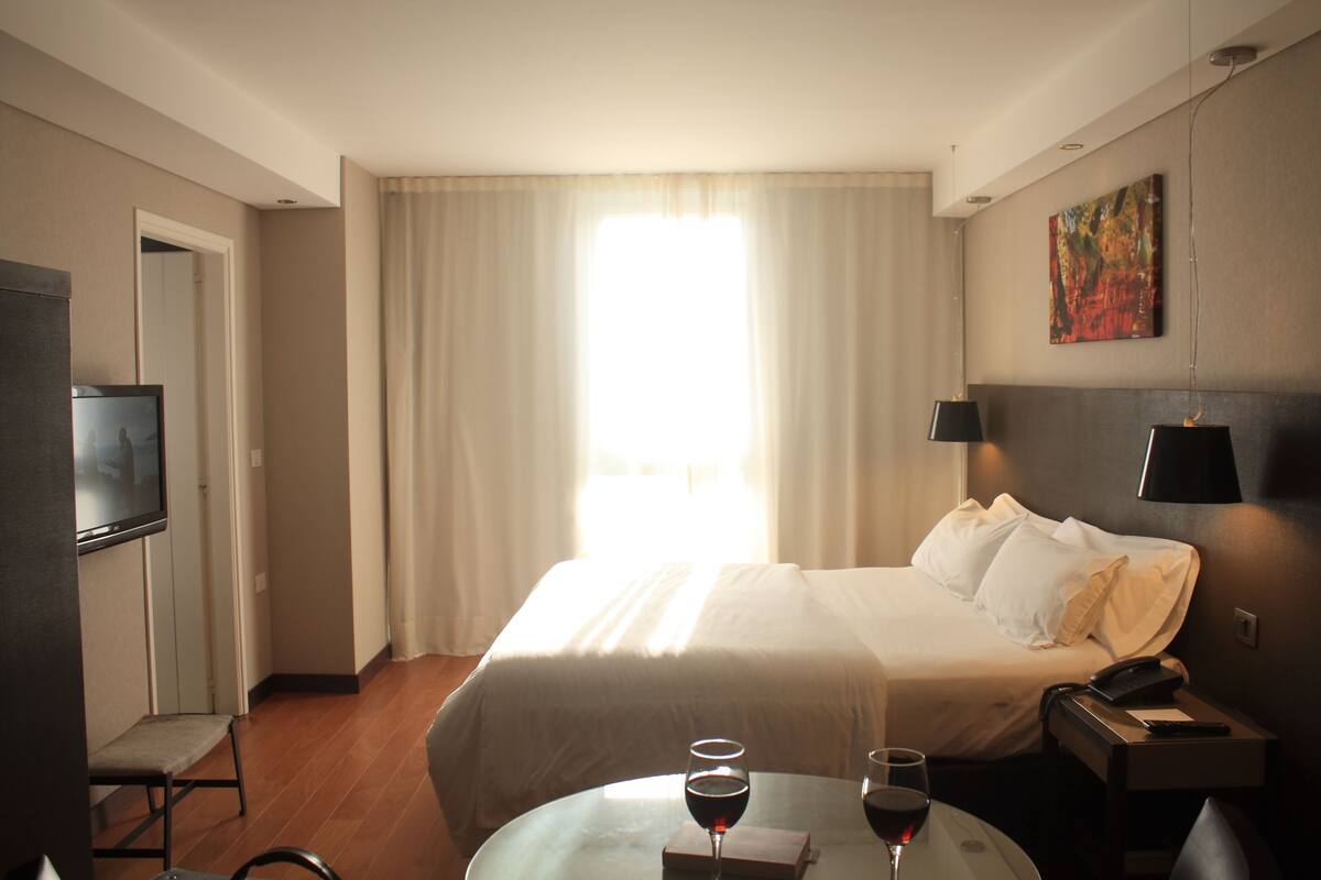 Habitación de hotel con una cama, un televisor, una mesa con dos copas de vino y un cuadro en la pared.