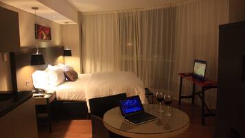 A hotel room with a bed, a desk with a laptop, a glass table with a wine glass, and a TV on a stand.