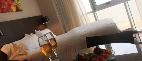 Habitación de hotel con una cama, una mesita de noche con una copa de vino, una bandeja de fruta y un cuadro en la pared.