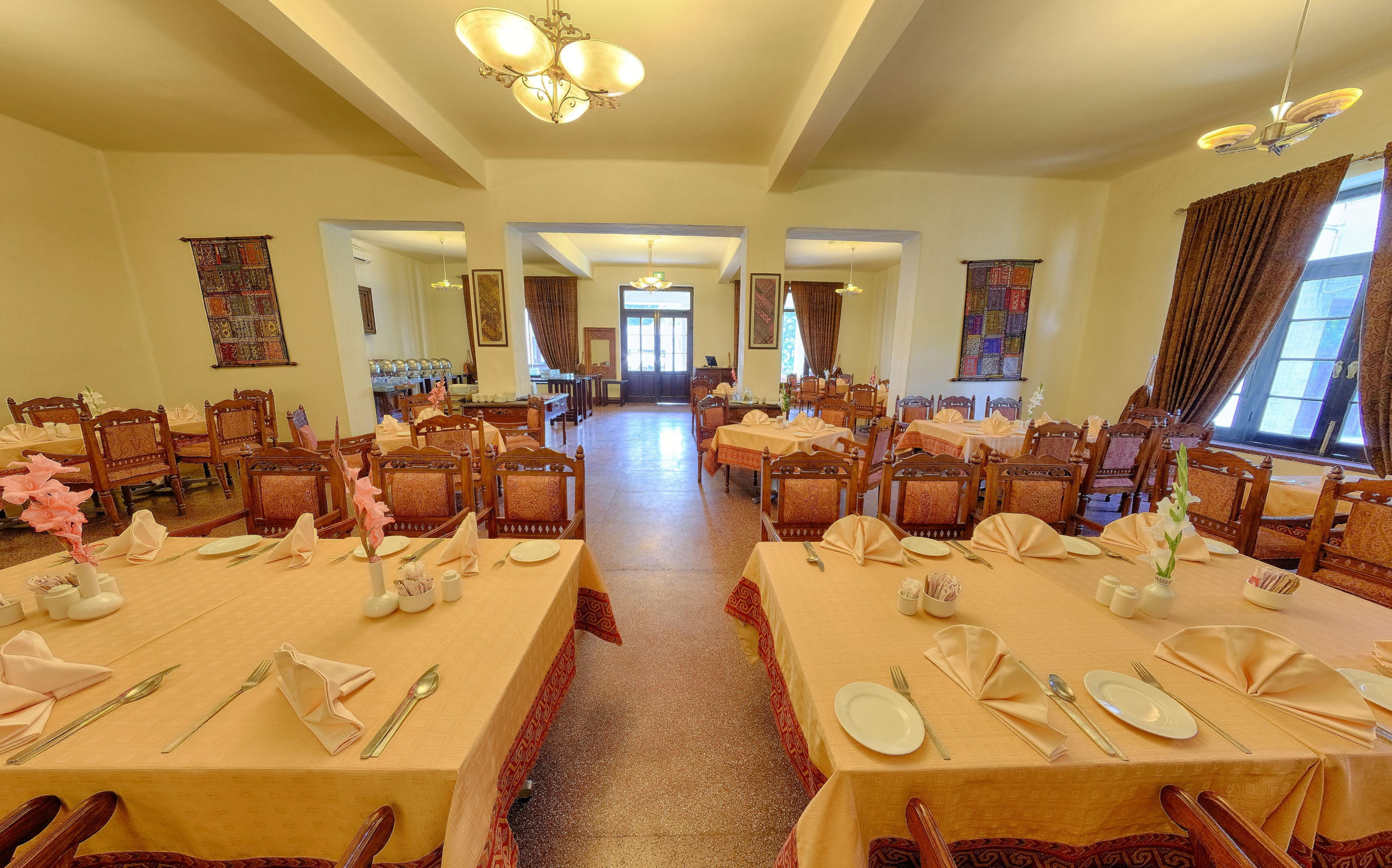 A spacious dining hall with multiple tables set for dining, featuring traditional wooden chairs and a central chandelier.