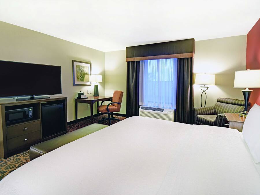 A hotel room with a large bed, a desk with a flat-screen TV, a chair, a sofa, and a window with curtains.