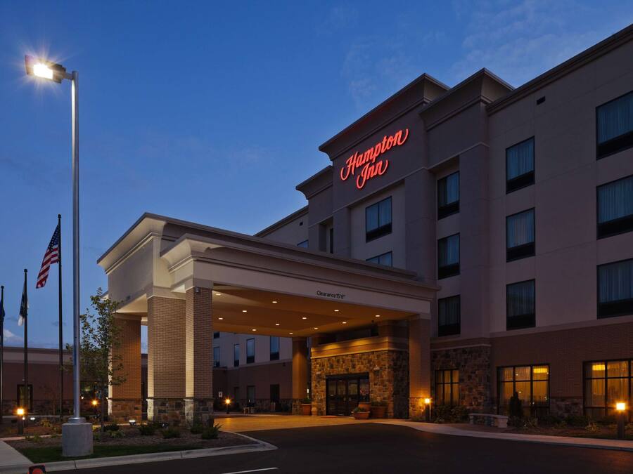 A Hampton Inn hotel entrance with a clear sky, American flags, and a well-lit facade.