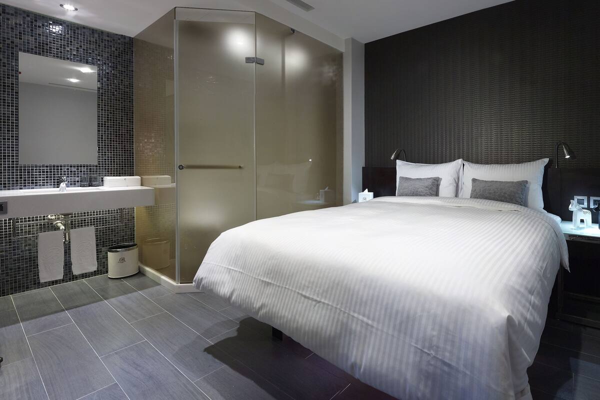 Moderni hotellihuone, jossa on suuri sänky, erillinen suihku, peilillä varustettu meikkipöytä ja tumma tehosteseinä.