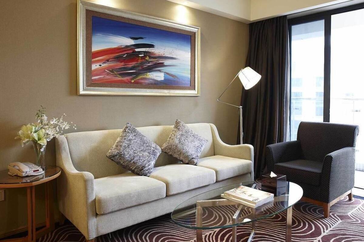 A modern hotel room with a beige sofa, a dark armchair, a glass coffee table, a painting on the wall, and a large window with curtains.