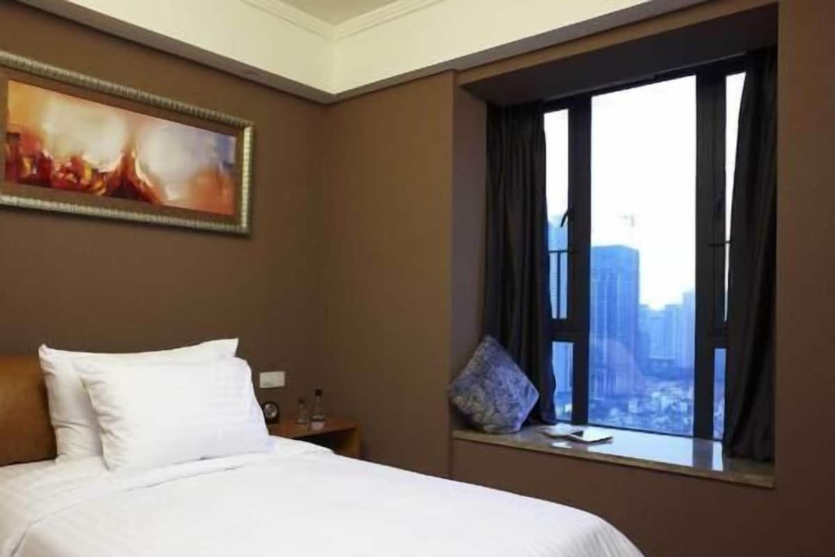 A hotel room with a bed, a window with a city view, a painting on the wall, and a nightstand with a lamp.