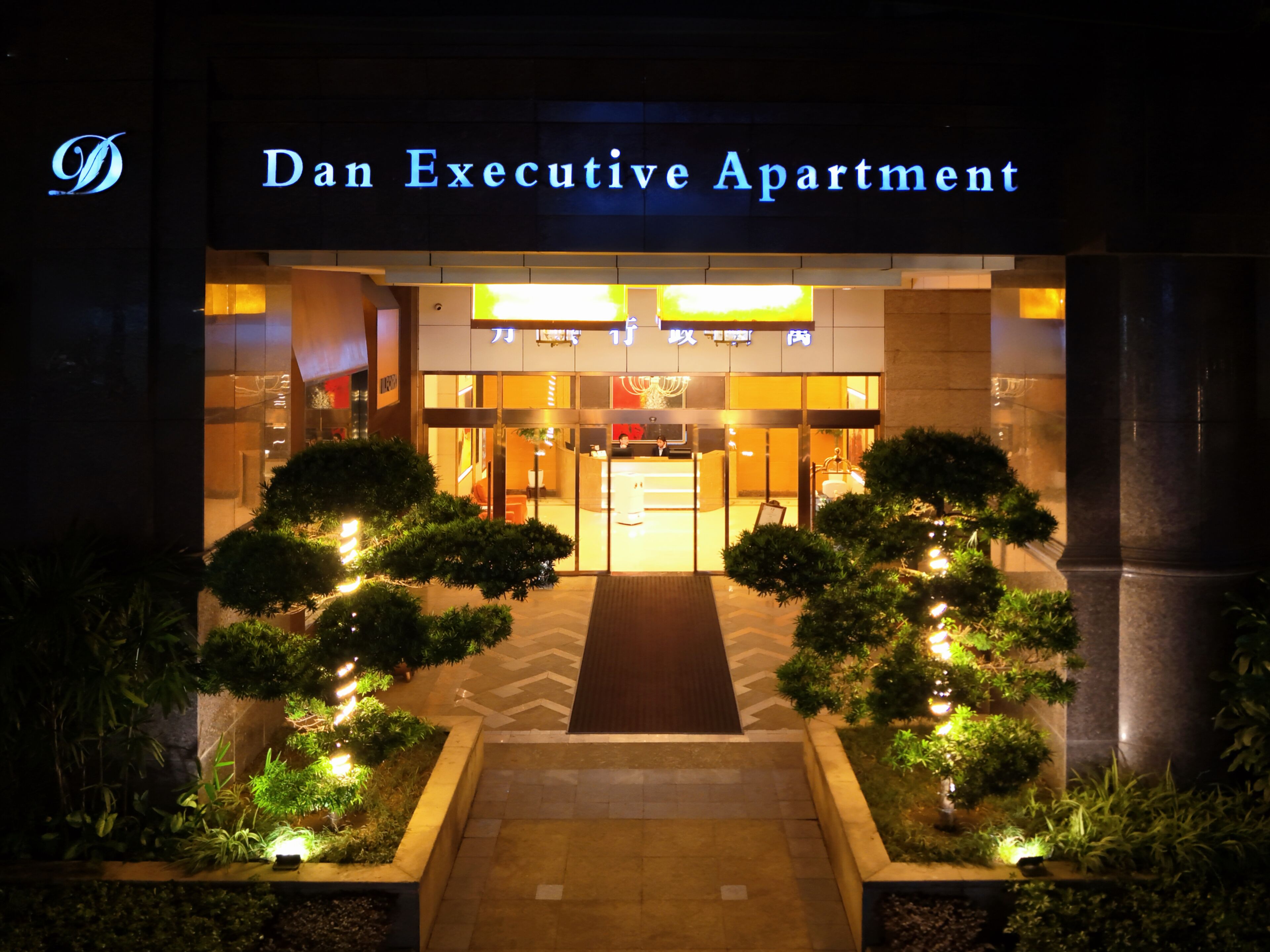 Indgangen til Dan Executive Apartment med en velbelyst facade, en central indgang og symmetrisk beplantning.