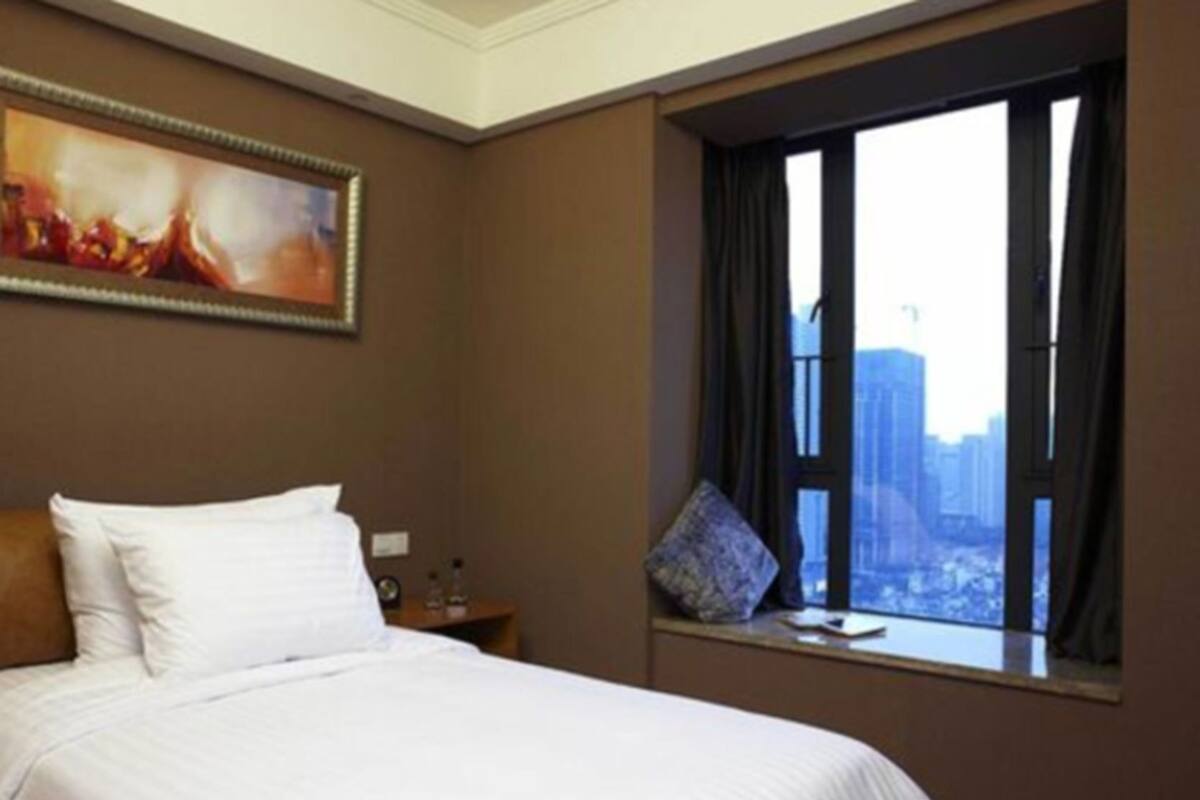 A hotel room with a large bed, a window with a city view, a painting on the wall, and a nightstand with a lamp.