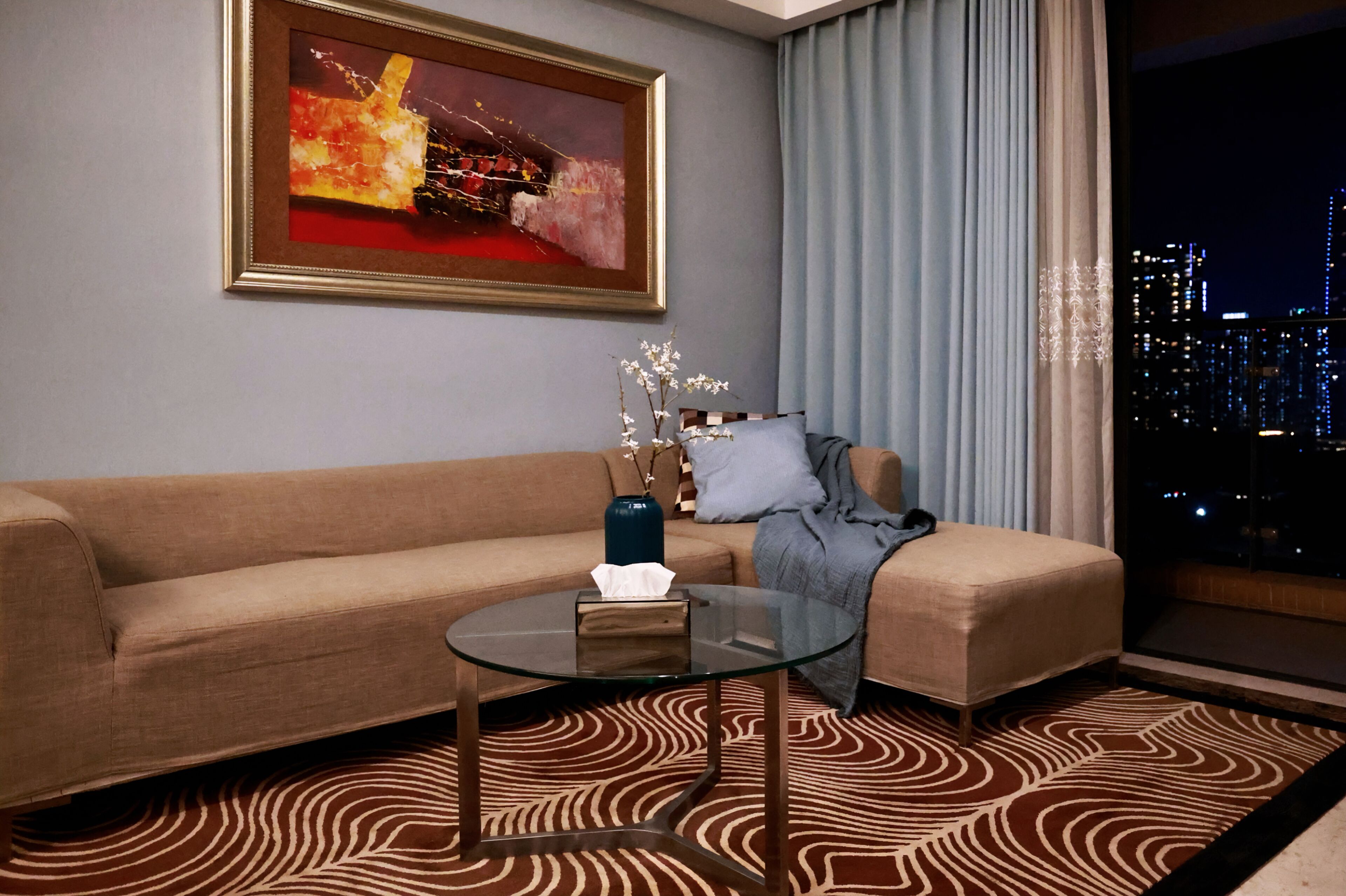 A modern living room with a sofa, a glass coffee table, a painting, a vase with branches, and a view of the city at night.