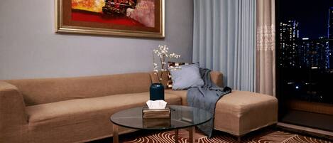 A modern living room with a sofa, a glass coffee table, a painting, a vase with branches, and a view of the city at night.