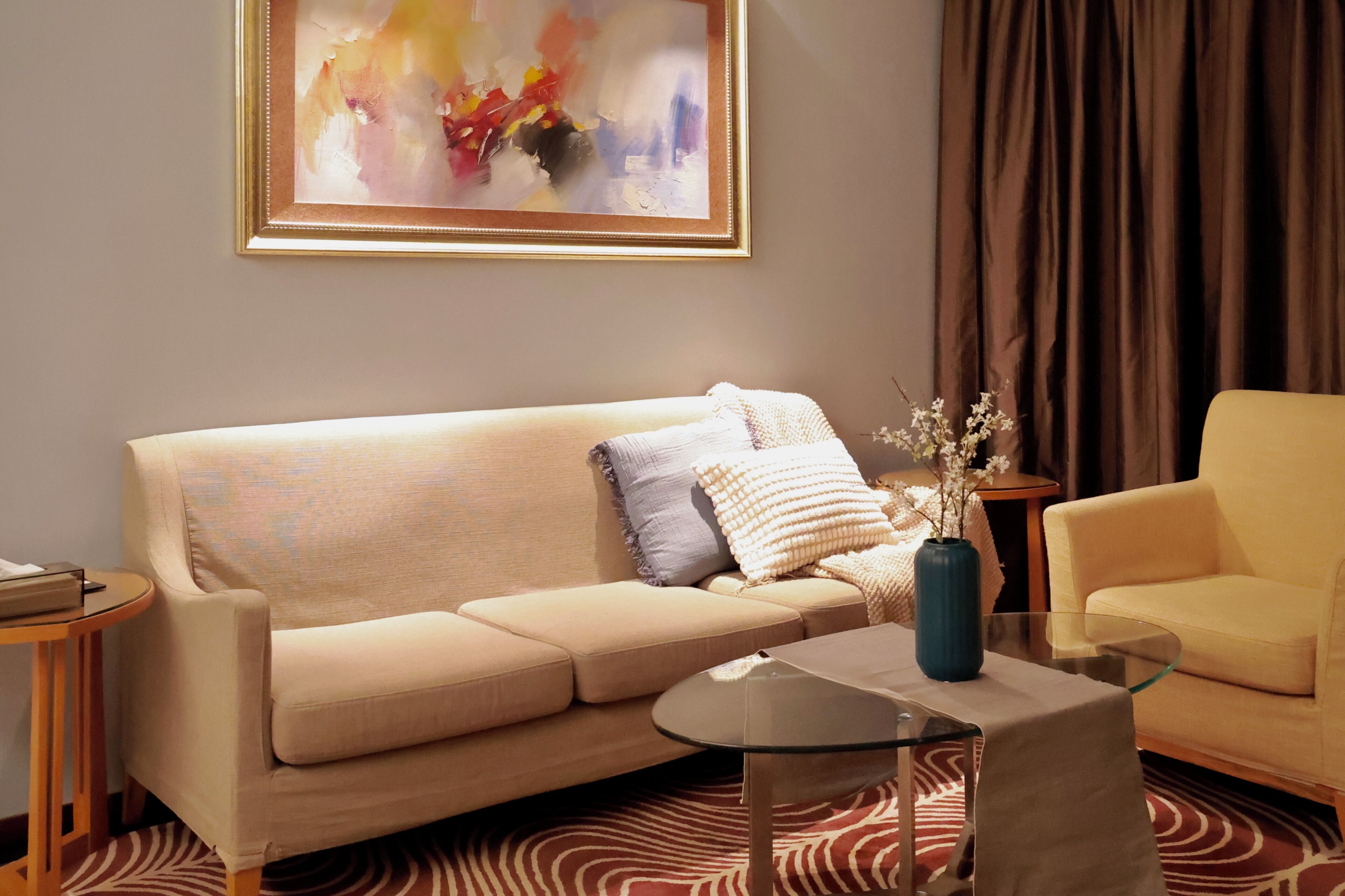 A living room with a beige sofa, a glass coffee table, a blue vase with flowers, a yellow armchair, and a framed abstract painting on the wall.