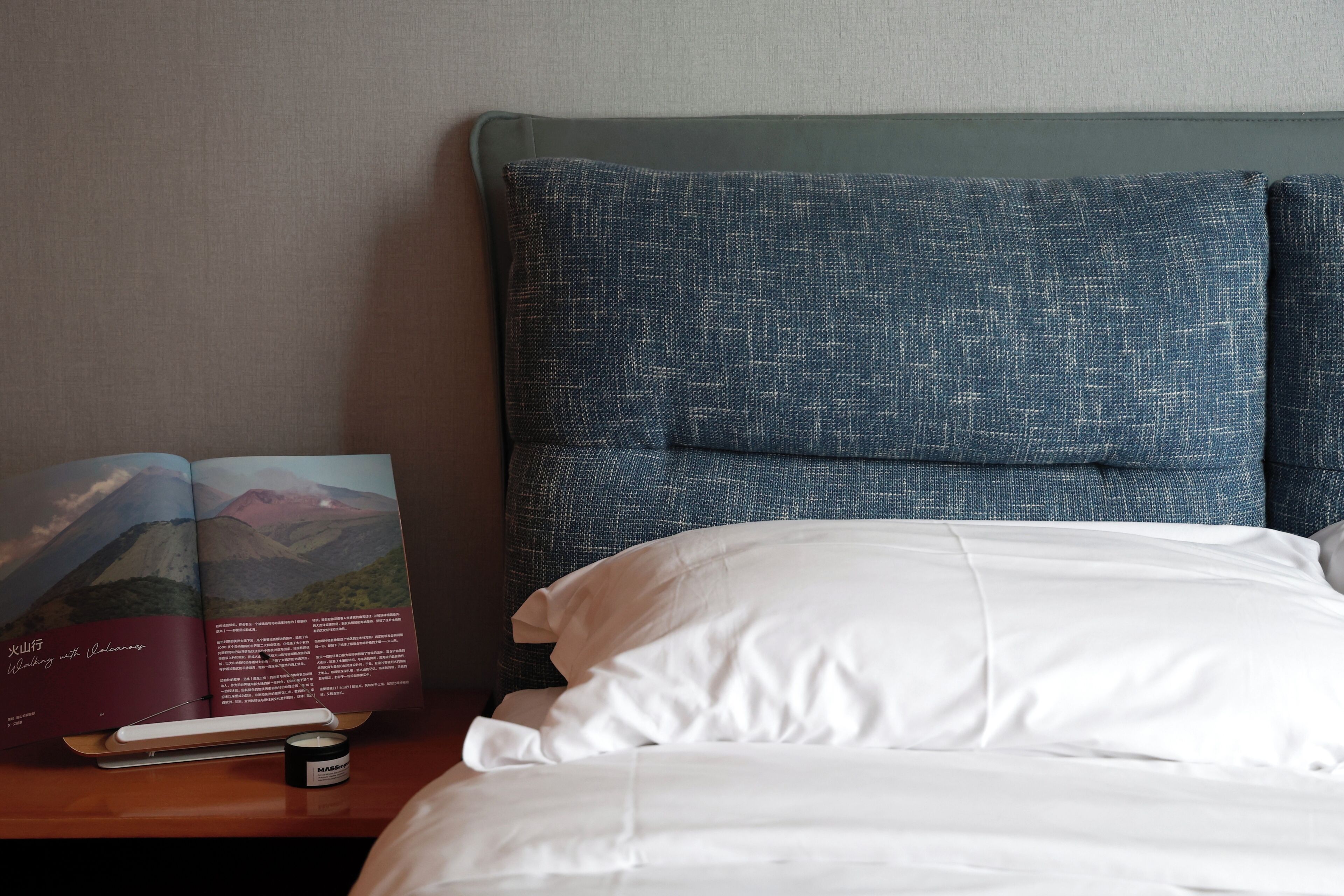 A neatly made bed with white linens, a book on a bedside table, and a small container of cream.