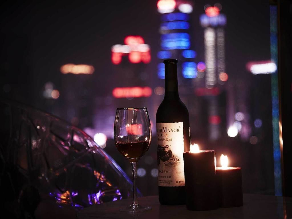 A bottle of wine and a glass on a table with a lit candle, set against a cityscape background.