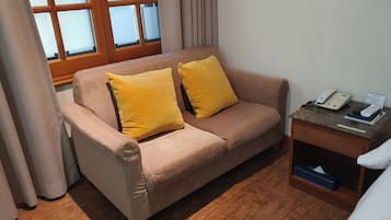 Ein Zimmer mit einem beigefarbenen Sofa, zwei gelben Kissen, einem Nachttisch aus Holz mit einem Telefon und einem Fenster mit Vorhängen.