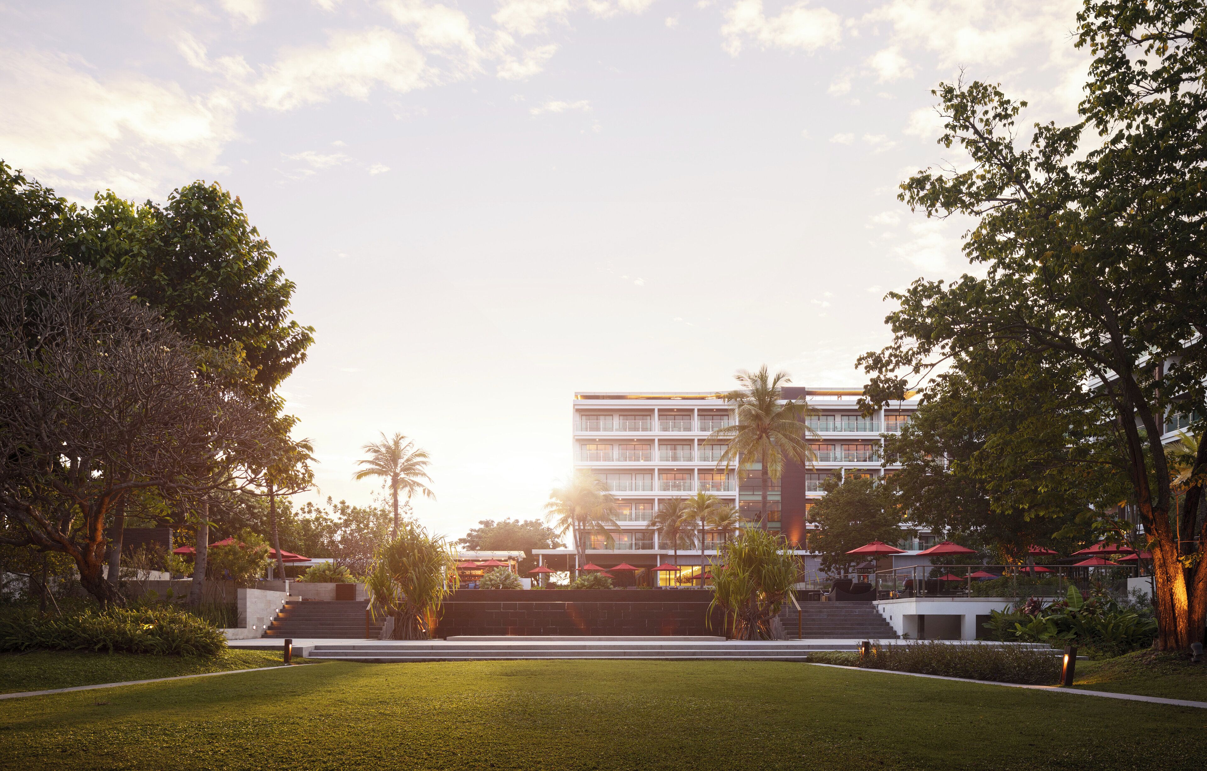 A hotel with a modern design, featuring a central staircase leading to the entrance, surrounded by lush greenery and palm trees.