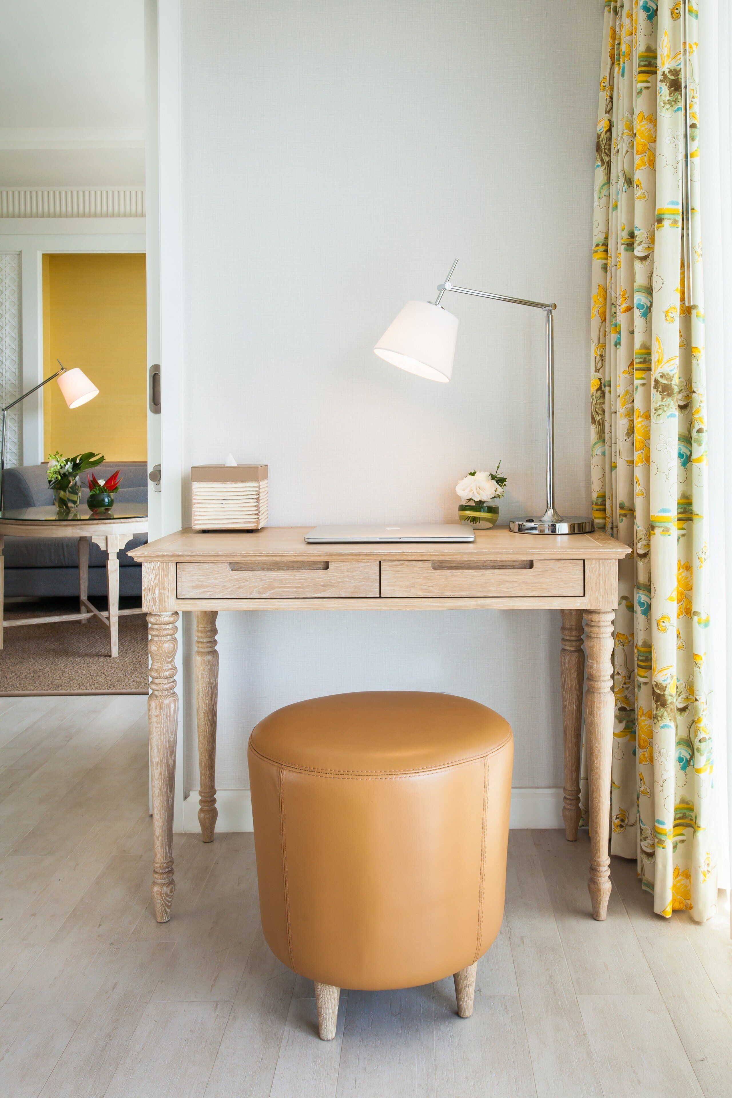 A room with a wooden desk, a laptop, a lamp, a small plant, and a leather ottoman.