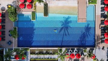 Vista aérea de un área de piscina de hotel con múltiples sombrillas rojas, sillas de descanso y palmeras.