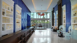 Un vestíbulo espacioso con un mostrador de recepción alargado, estanterías blancas y azules que exhiben diversos objetos decorativos, y un amplio ventanal con vistas al exterior.