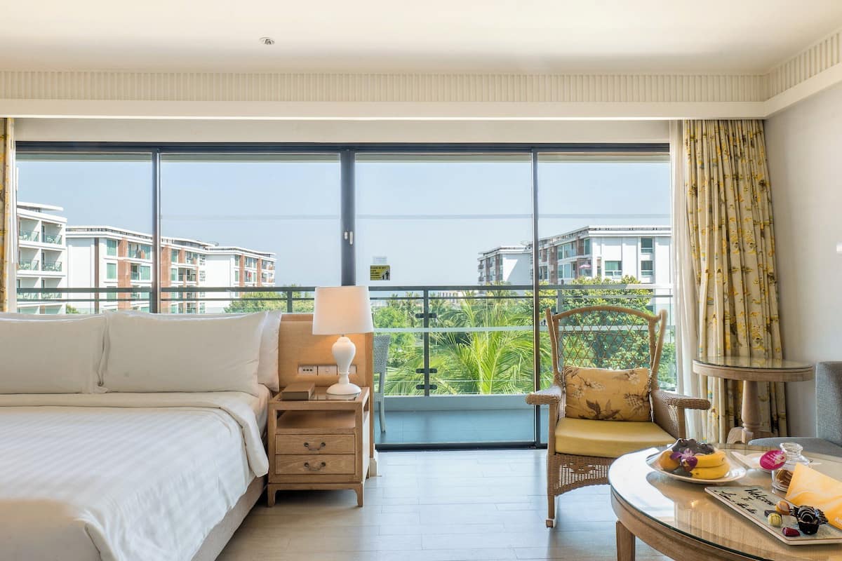 Habitación de hotel con una cama grande, una mesita de noche, una silla, una mesa con una bandeja de comida y vistas a edificios y vegetación a través de amplios ventanales.