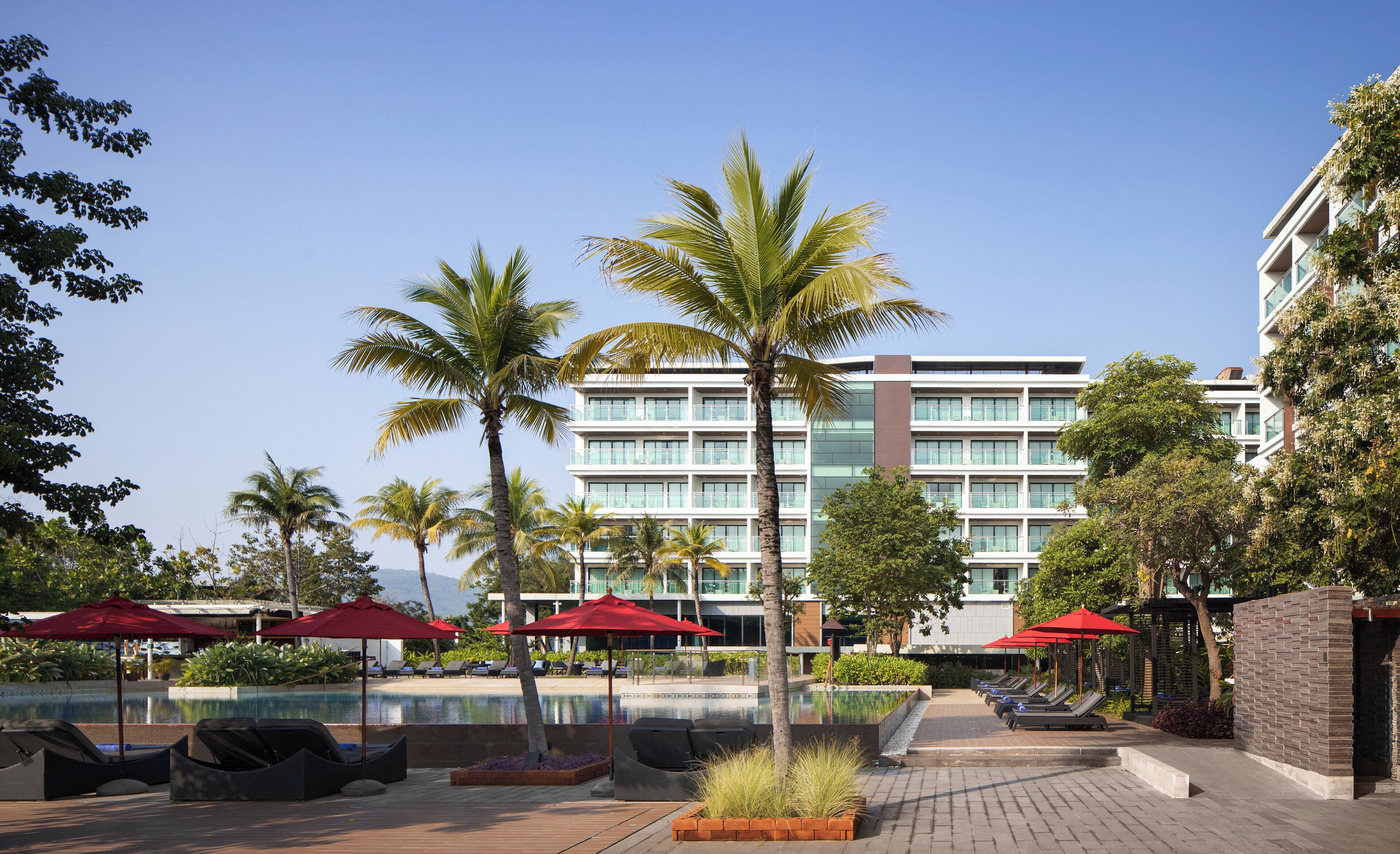 Un hotel con piscina, palmeras y sombrillas rojas.