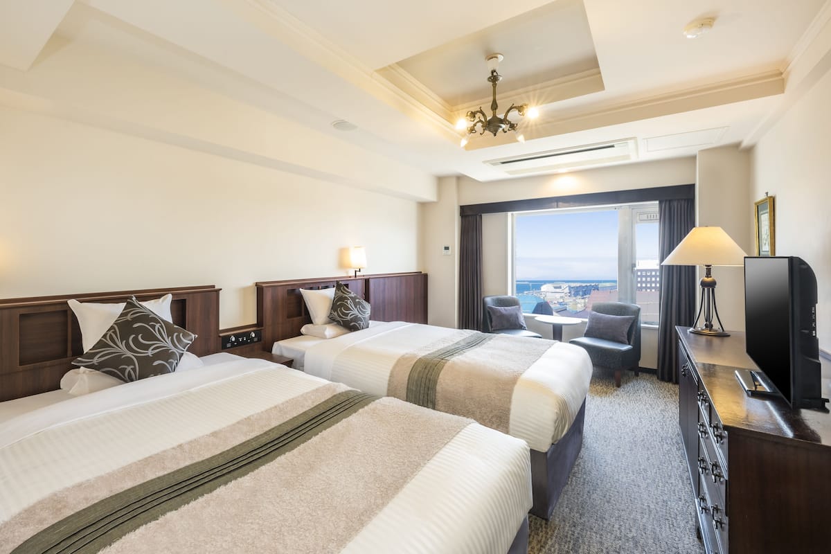A hotel room with two beds, a desk with a lamp, a TV, and a view of the city.