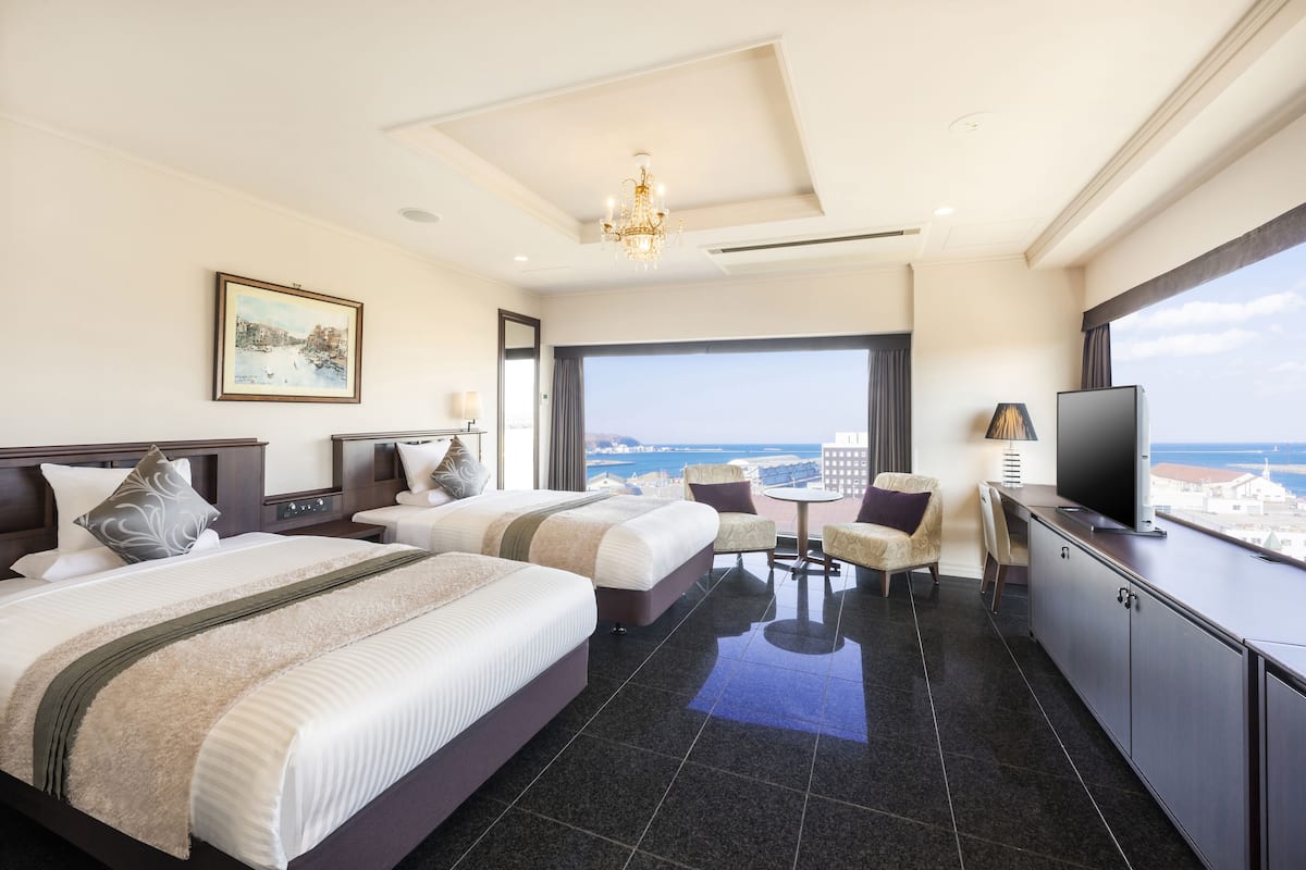 A hotel room with two beds, a TV, a desk, and a view of the sea.