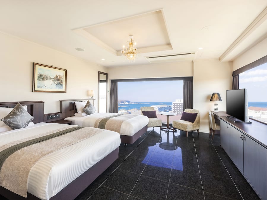 A hotel room with two beds, a TV, a desk, and a view of the sea.